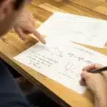 Tutor mengarahkan anak mengerjakan lembar kerja matematika di meja rumah